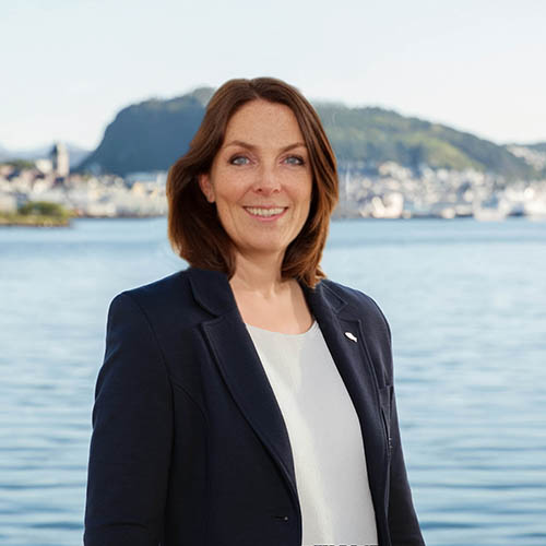 Smiling woman with shoulder-length brown hair wearing a navy blazer and white top, standing in front of a calm body of water with a blurry mountainous town background.