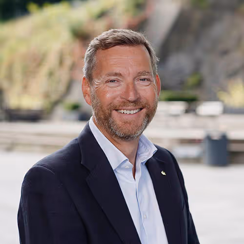Smiling middle-aged man with light brown hair and beard wearing a dark blazer and white shirt outdoors.