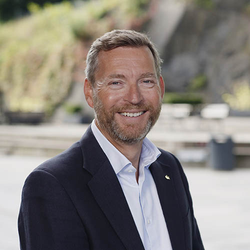 Smiling middle-aged man with light brown hair and beard wearing a dark blazer and white shirt outdoors.