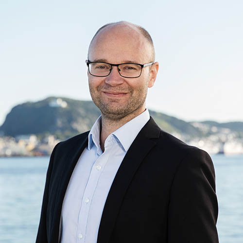 Man with glasses wearing a light blue shirt and black jacket, standing in front of a waterfront and distant hills.