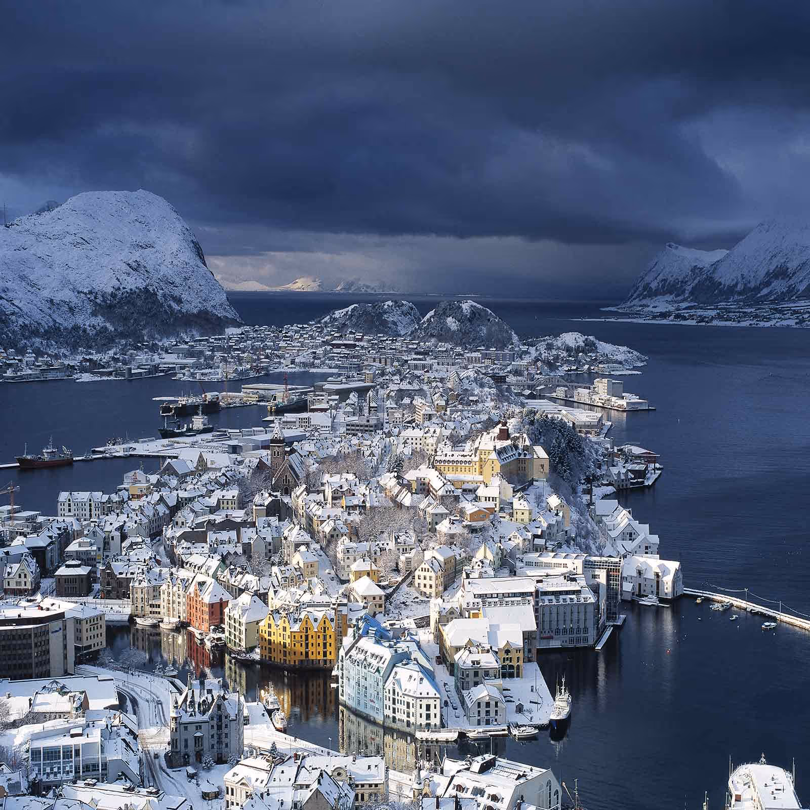 Aalesund town covered in snow with colorful buildings by the water and snow-covered mountains under a dark cloudy sky.