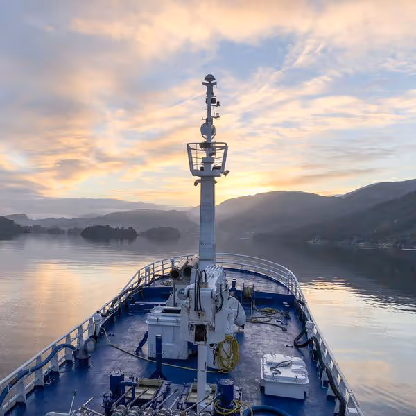 View from the bow of a ship on calm water with a mountainous coastline and a colorful sunset sky.