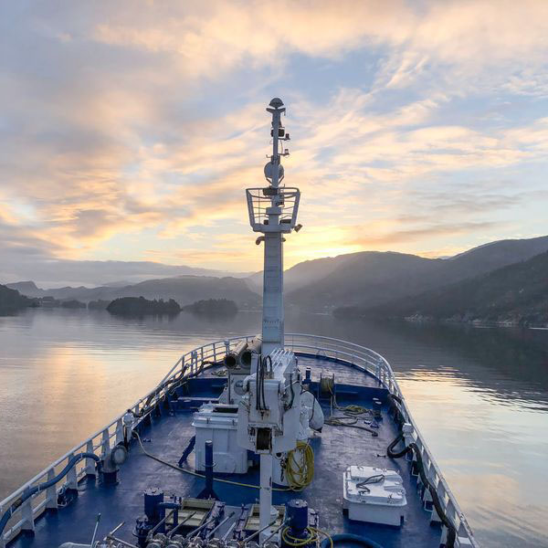View from the bow of a ship on calm water with a mountainous coastline and a colorful sunset sky.