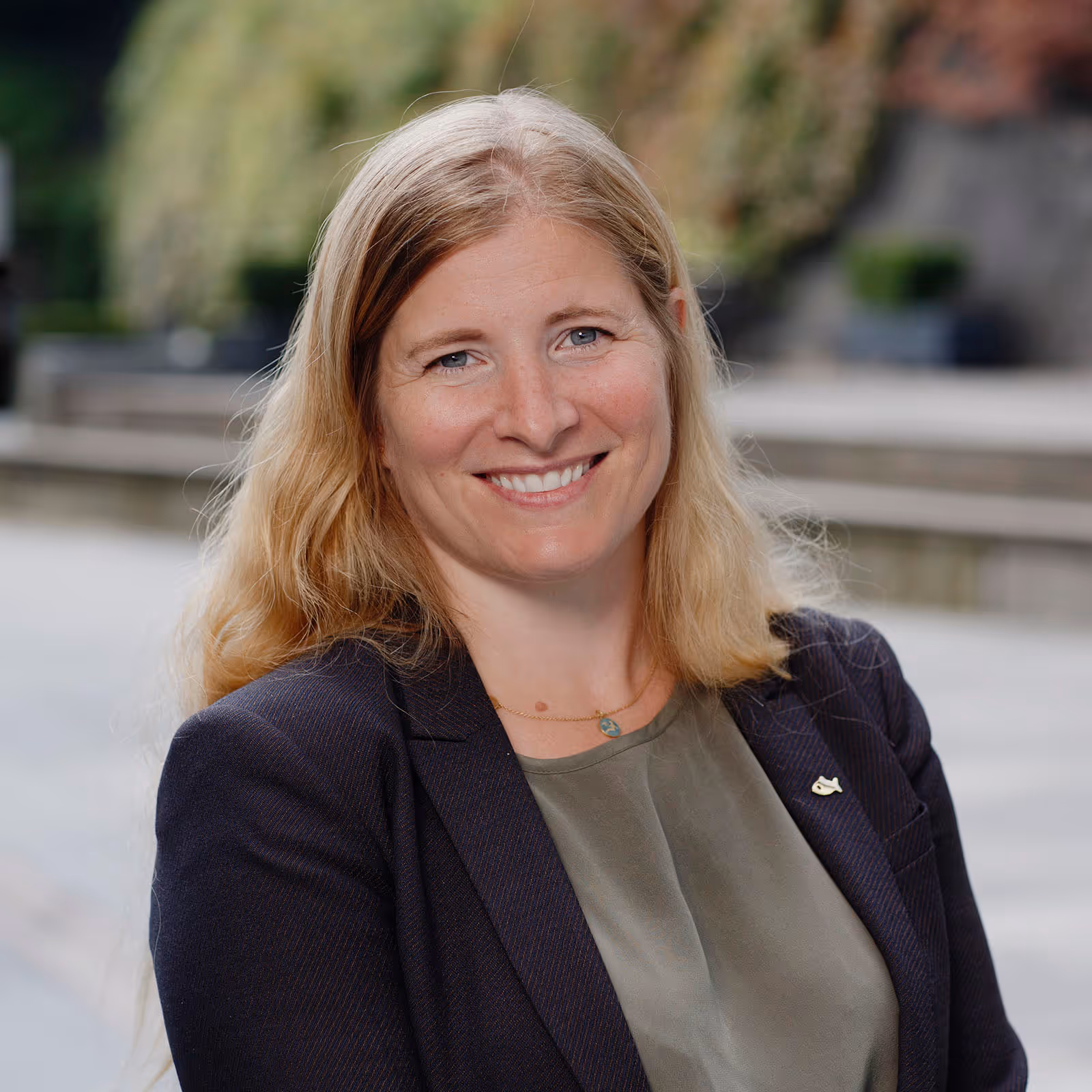 Smiling woman with long blonde hair wearing a dark blazer and olive green top outdoors.