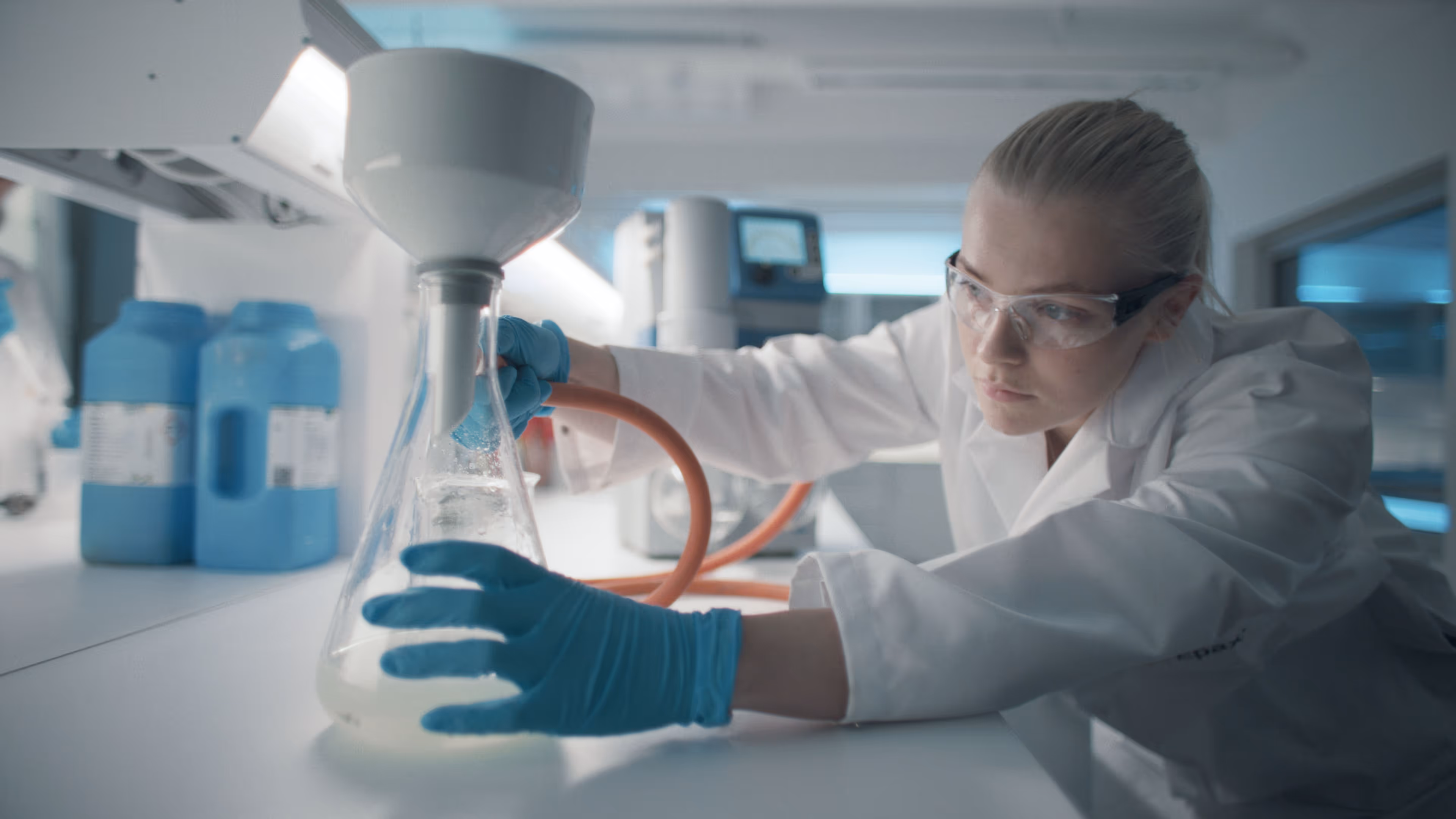 Female scientist in lab coat and safety glasses handling a conical flask with liquid under a lab funnel.