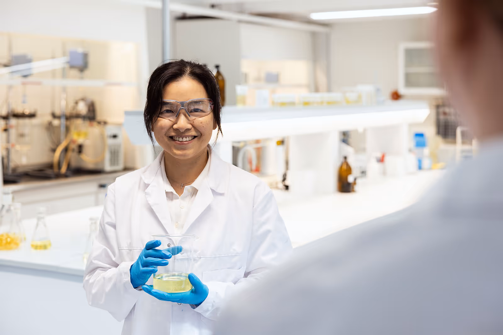 Female scientist wearing safety glasses and blue gloves holding a glass container with yellow liquid in a laboratory.