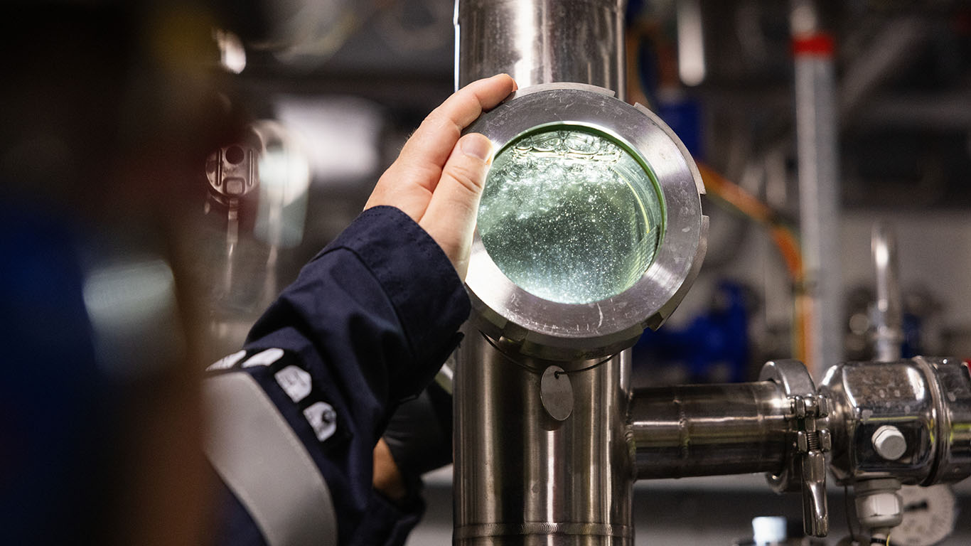 Hand inspecting bubbles and liquid flow through a round glass window on a stainless steel industrial pipe system.