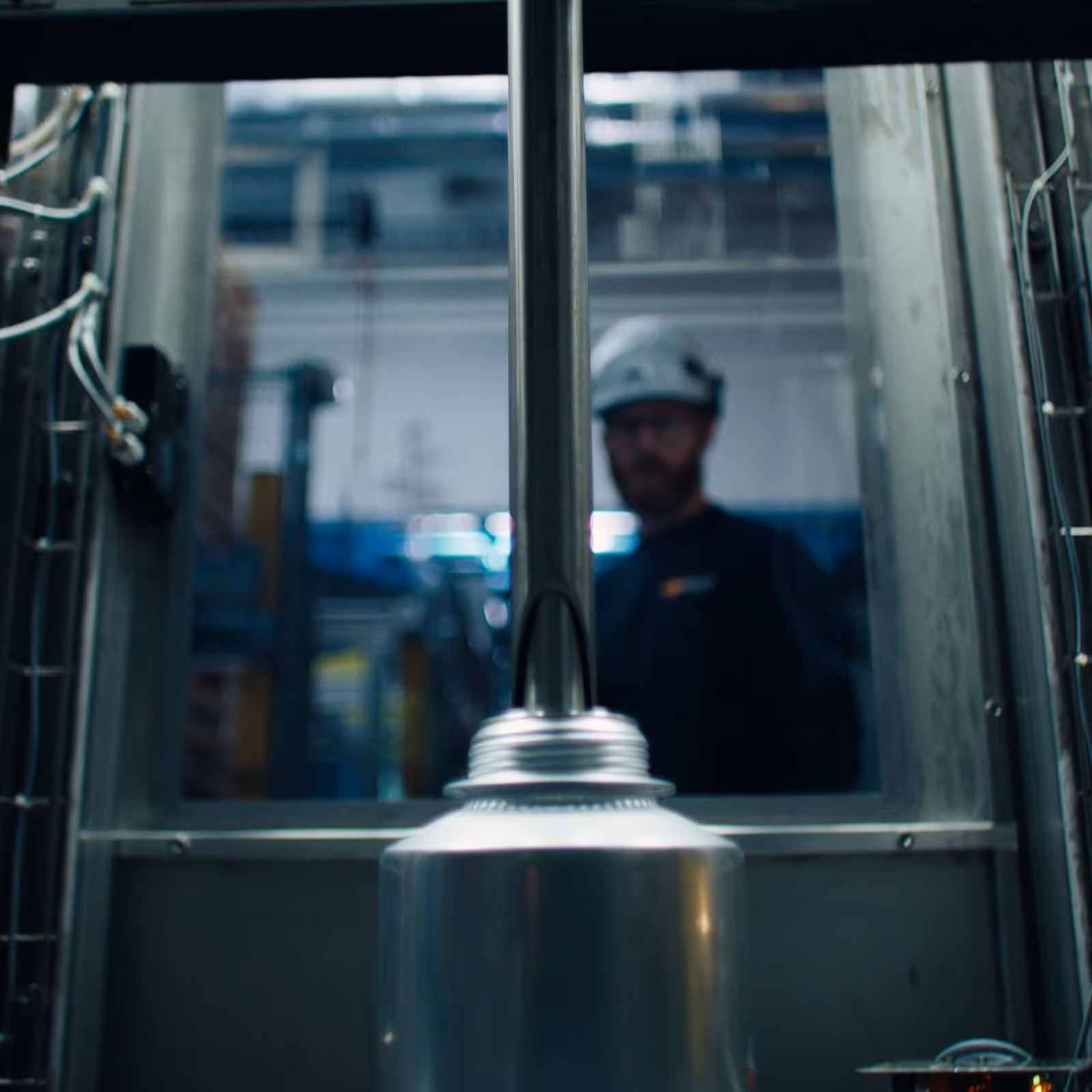 Close-up of industrial equipment with a blurred worker wearing a white hard hat in the background.