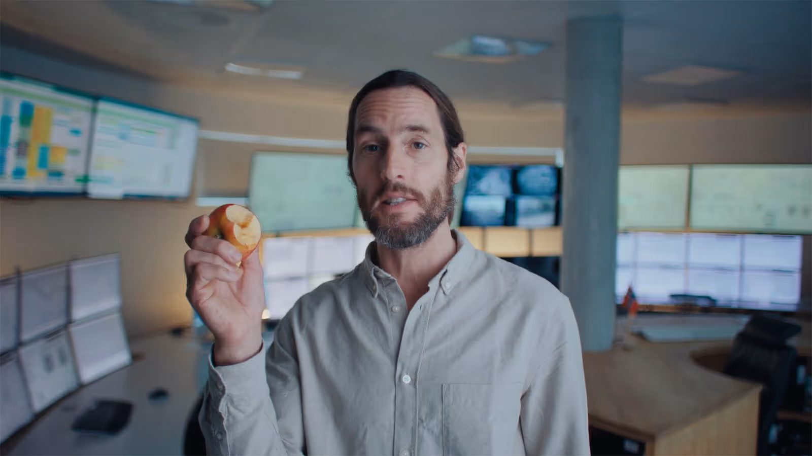 Man with beard holding a partially eaten apple in an office with multiple computer monitors.