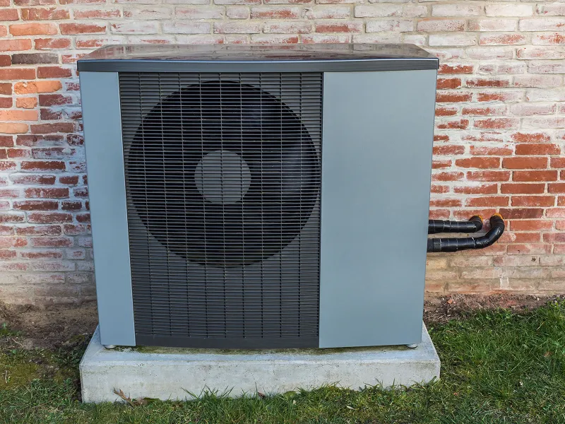 Außenstehende Wärmepumpe mit schwarzen Rohren an einer Backsteinmauer.