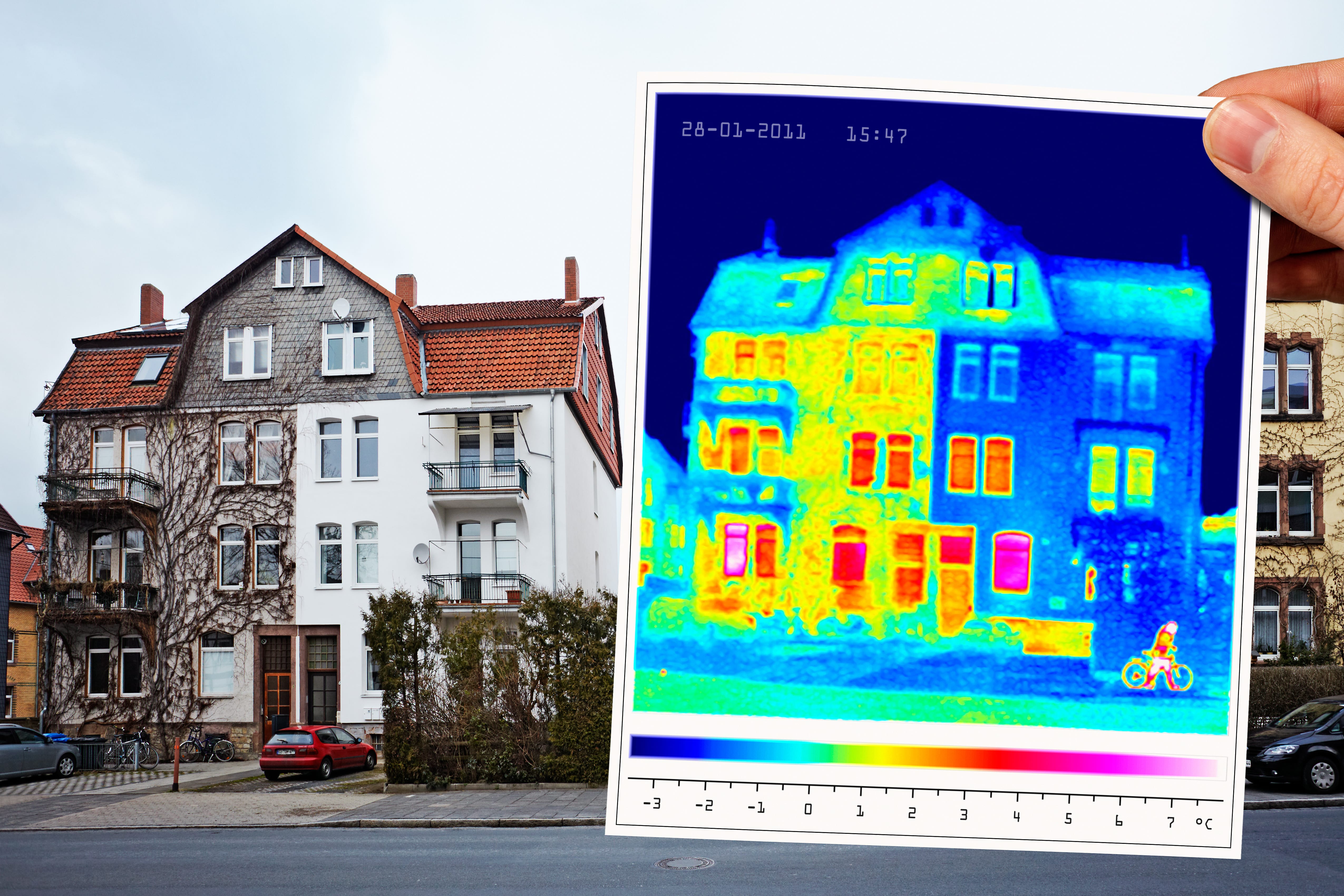 Eine Person hält eine Thermografieaufnahme eines Hauses, die Wärmeverluste in Gelb und Rot anzeigt, daneben das reale Haus.