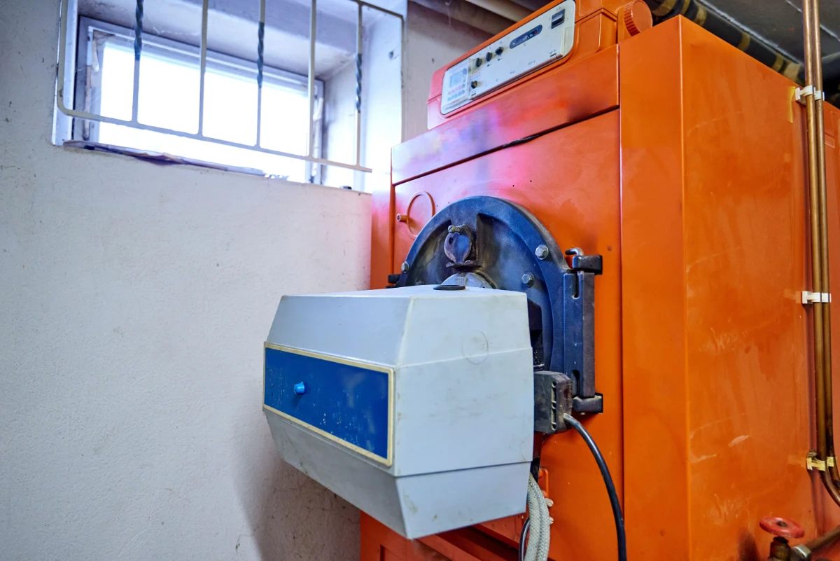 Alter großer oranger Ölkessel mit weiß-blauem Bedienfeld in einem Kellerraum unter einem vergitterten Fenster.