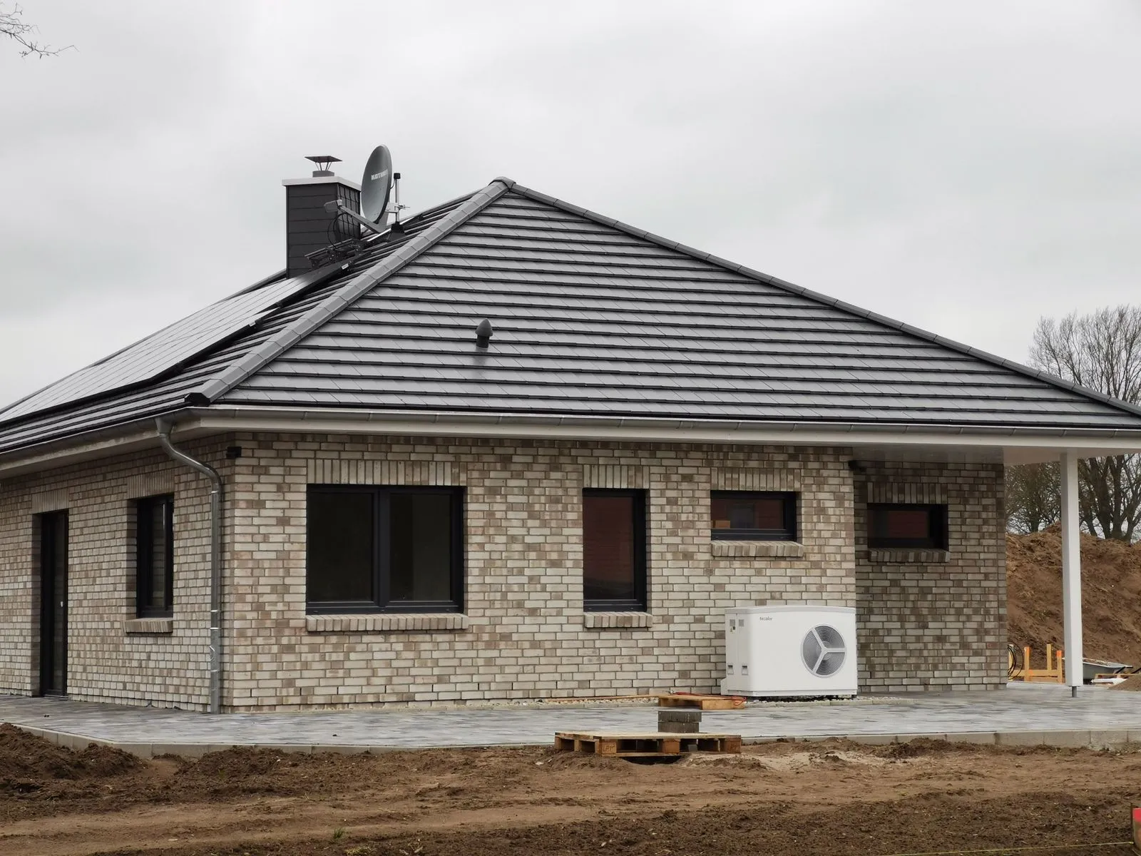 Ein niedriger Neubau mit grauem Ziegeldach, Solarpanelen auf der linken Seite und einer installierten Wärmepumpe vor dem Haus.