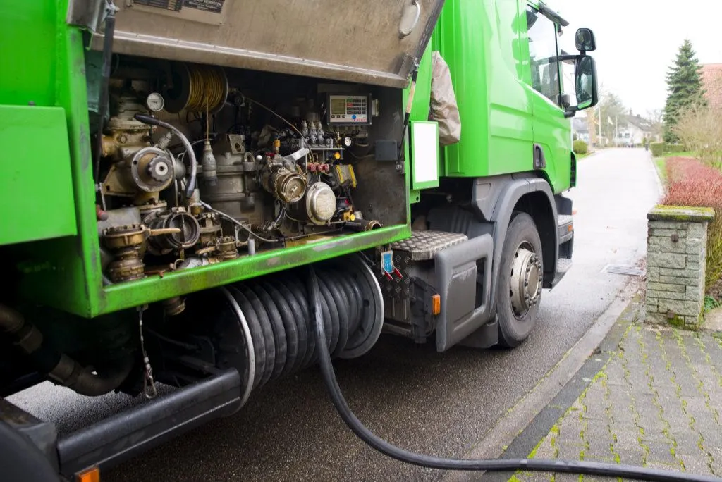 Grüner LKW mit offener Seitenklappe, die Heizöllieferungsausrüstung und einen schwarzen Schlauch zeigt, der auf einer nassen Straße liegt.