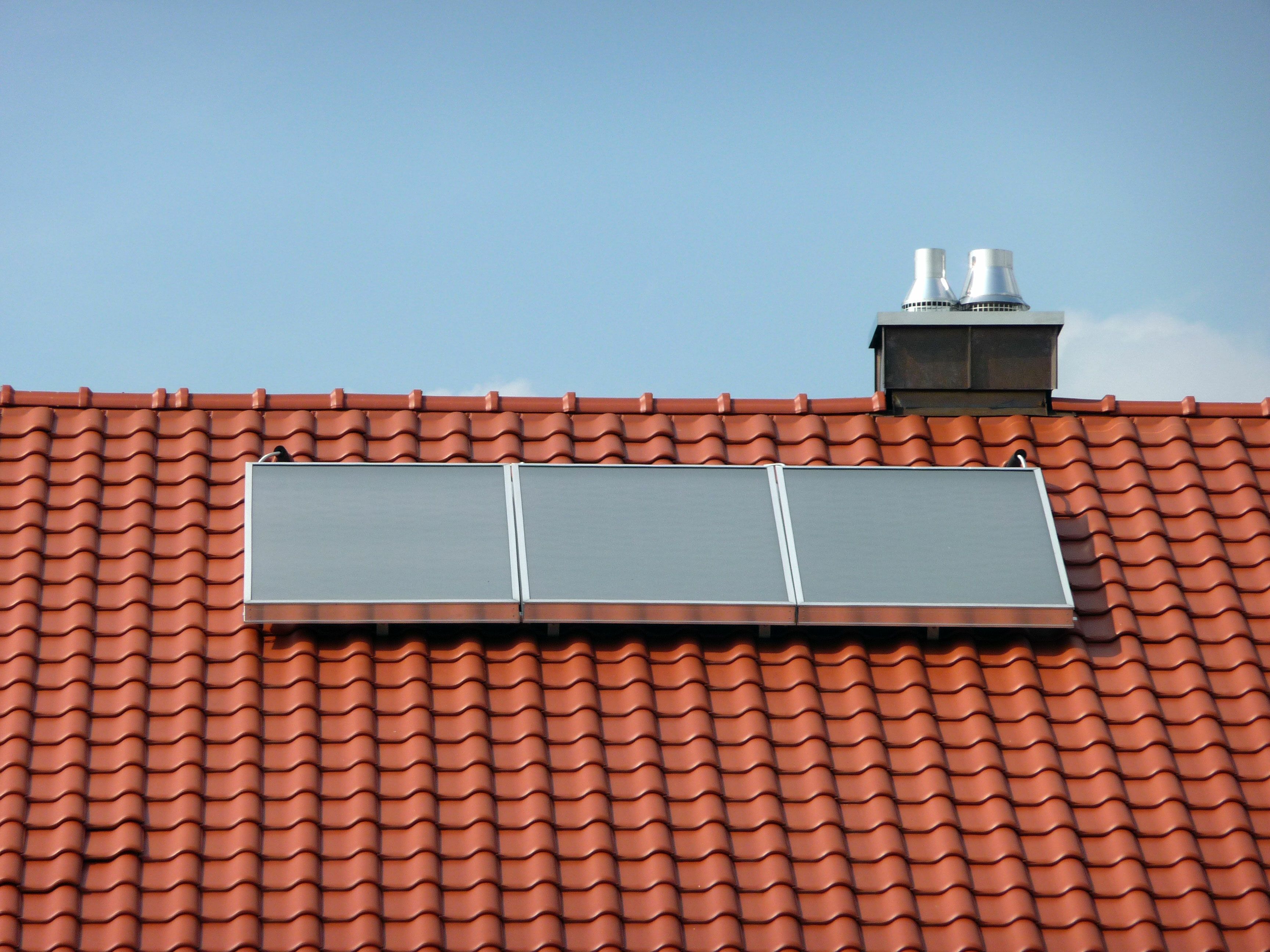 Ölheizung hybrid mit Solarthermie: Hausdach mit roten Ziegeln, drei Solarthermie-Kollektoren und Schornstein.