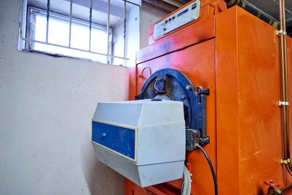 Orange Ölheizung in einem Kellerraum neben einem kleinen Gitterfenster.
