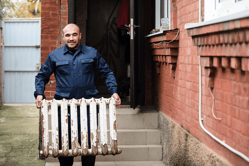 Mann in blauem Arbeitsanzug trägt einen alten, abgeblätterten weißen Heizkörper draußen vor einem roten Backsteinhaus.