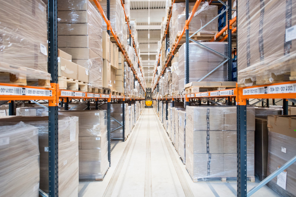Lagerhalle mit langen Regalgängen voller gestapelter und in Folie verpackter Kartons auf Paletten.