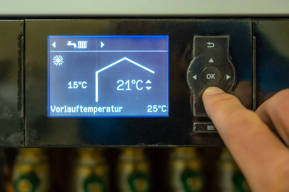 Hand stellt Vorlauftemperatur von 21 Grad Celsius an digitalem Heizungssteuergerät ein.