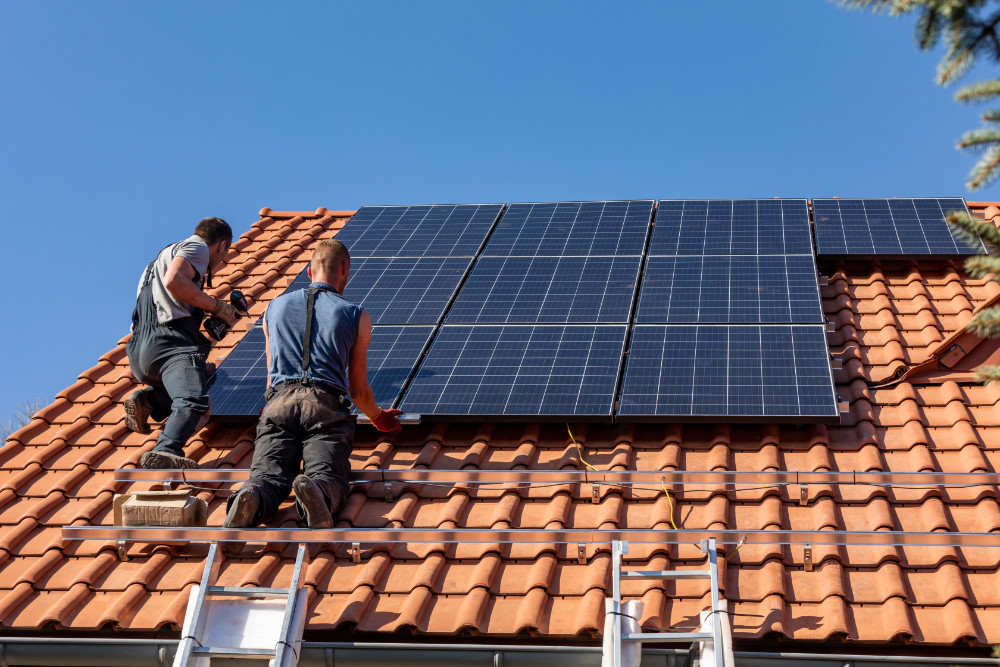 Zwei Handwerker montieren Solarpaneele auf einem Dach mit roten Ziegeln unter klarem blauem Himmel.