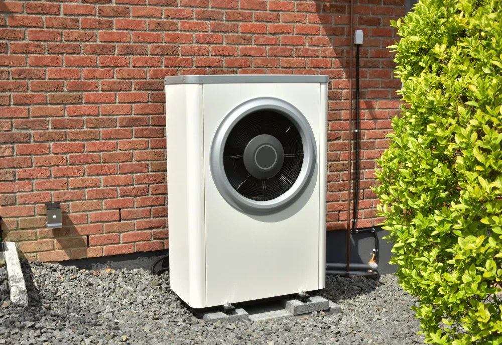 Weiße Außeneinheit einer Wärmepumpe vor roter Ziegelwand neben einem grünen Busch.