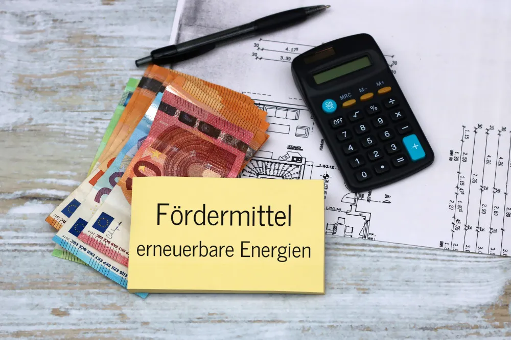 Mehrere Euro-Banknoten, ein Taschenrechner, ein Stift und ein Bauplan liegen auf einem Holztisch neben einem Zettel mit der Aufschrift »Fördermittel erneuerbare Energien«.