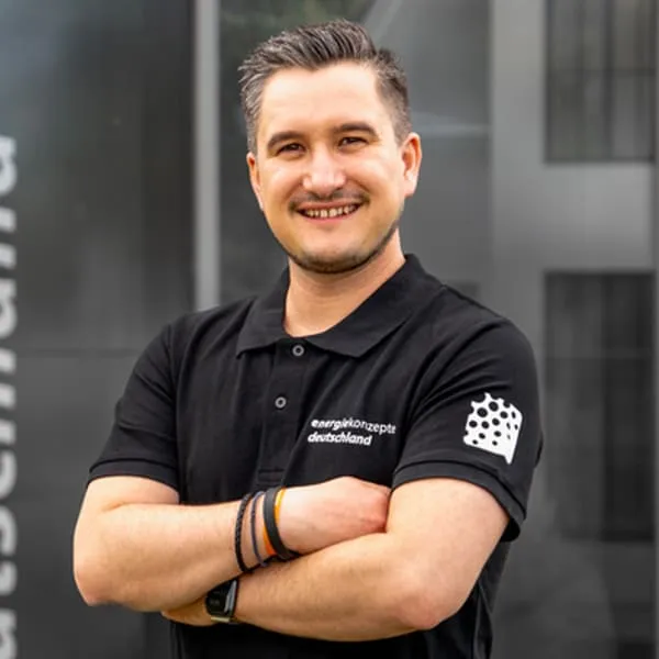 Junger Mann mit verschränkten Armen trägt schwarzes Poloshirt mit Aufschrift energiekonzepte deutschland vor modernem Gebäude.