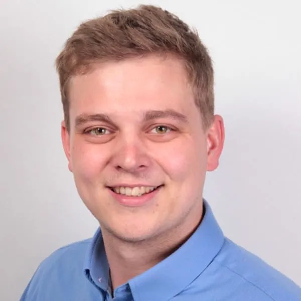 Junger Mann mit kurzem braunem Haar, lächelnd, trägt ein hellblaues Hemd vor weißem Hintergrund.