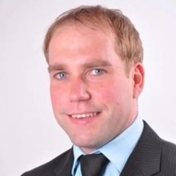 Porträt eines lächelnden Mannes mit kurzem blondem Haar, blauem Hemd, schwarzer Krawatte und dunklem Anzug.