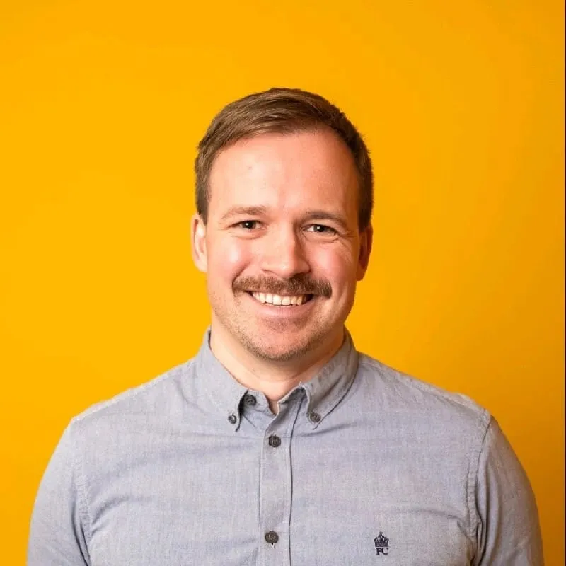 Porträt eines lächelnden Mannes mit Schnurrbart vor einem orangefarbenen Hintergrund, der ein hellblaues Hemd trägt.