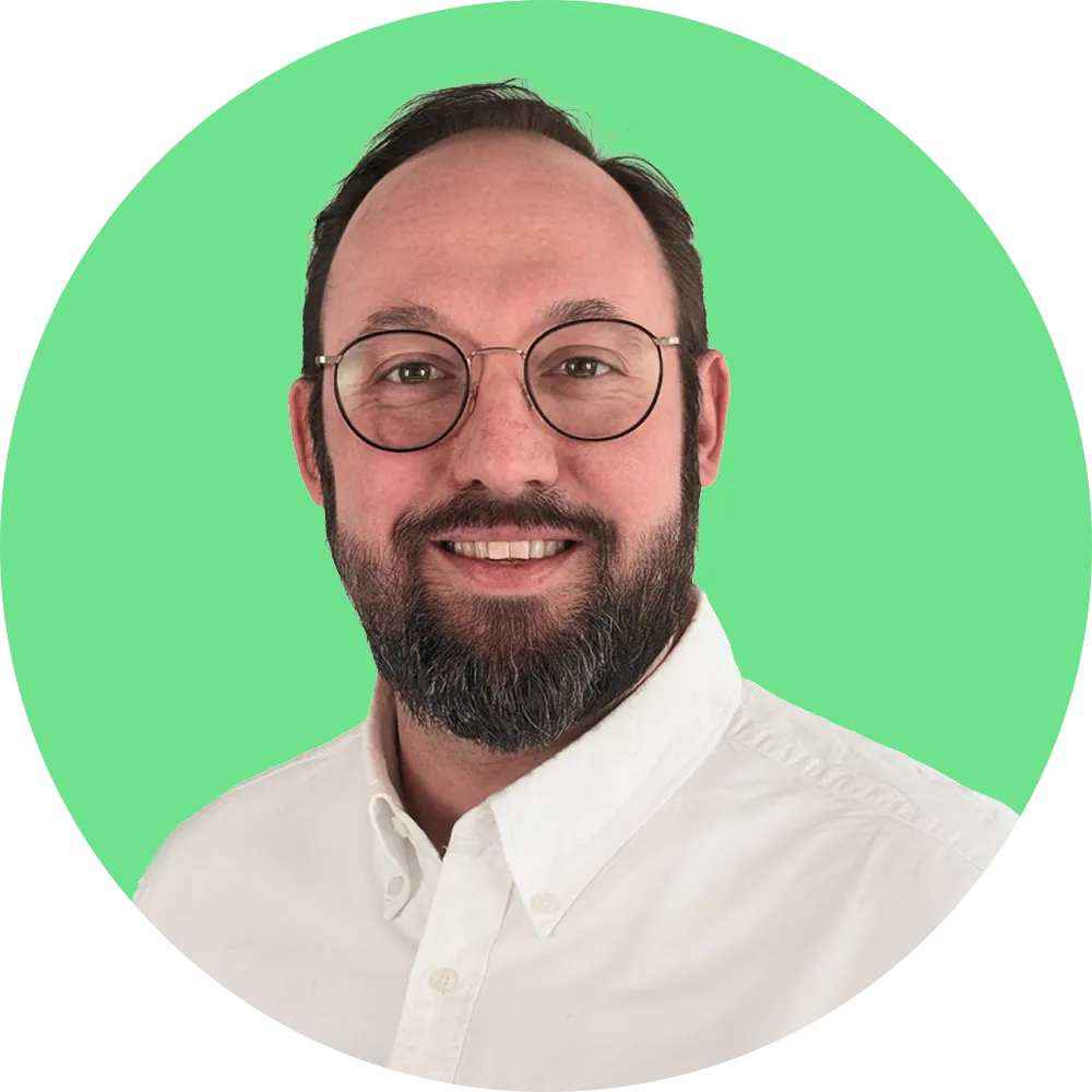 Porträt eines lächelnden Mannes mit Brille, Bart und weißem Hemd vor grünem Hintergrund.