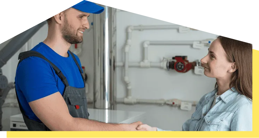 Ein Monteur in blauer Arbeitskleidung und eine Frau geben sich die Hand in einem Technikraum mit Rohren und Ventilen.