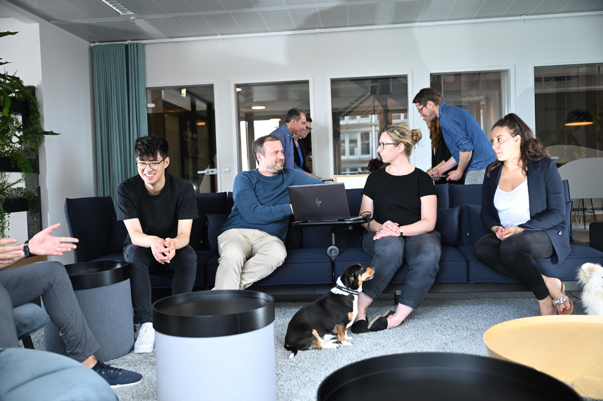 Eine Gruppe von Menschen in einem modernen Büro sitzt auf Sofas, darunter ein Mann mit einem Laptop und zwei Hunden auf dem Boden.