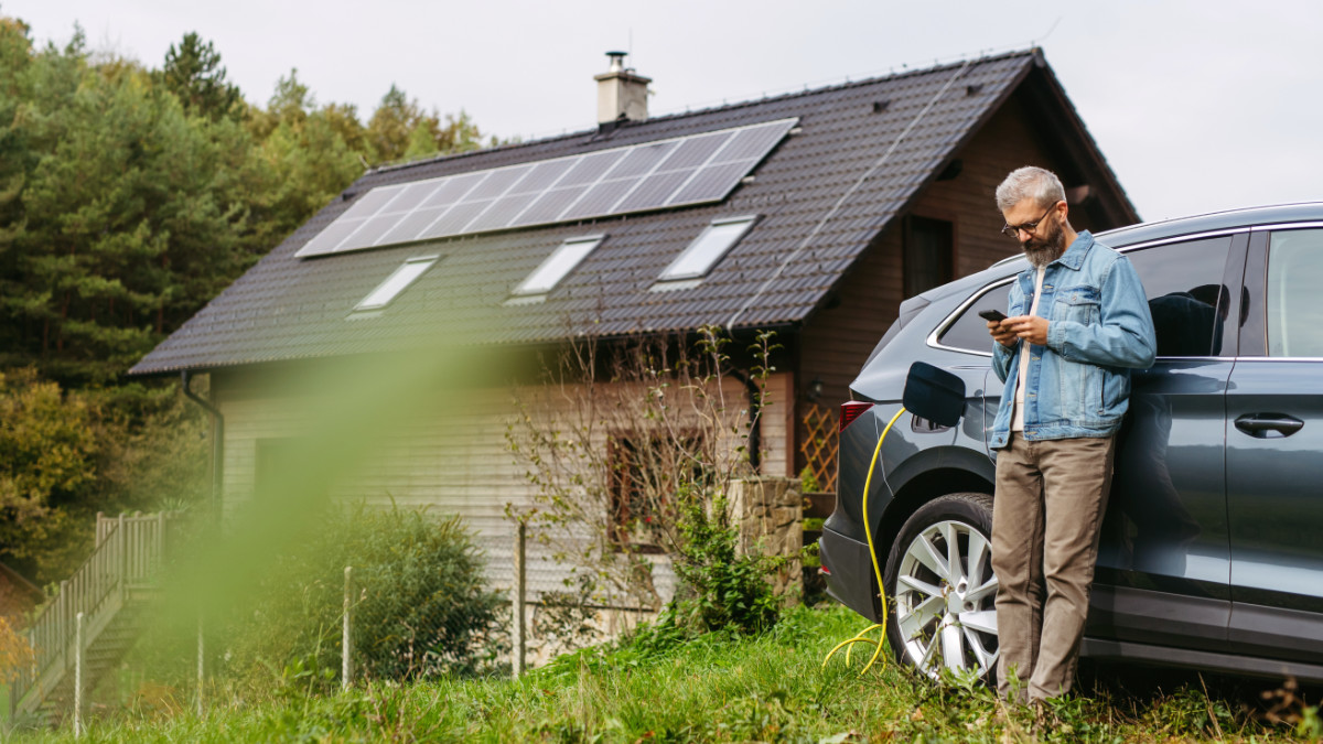 Mann steht neben einem Elektroauto, das vor einem Haus mit Solarpaneelen auf dem Dach geladen wird.