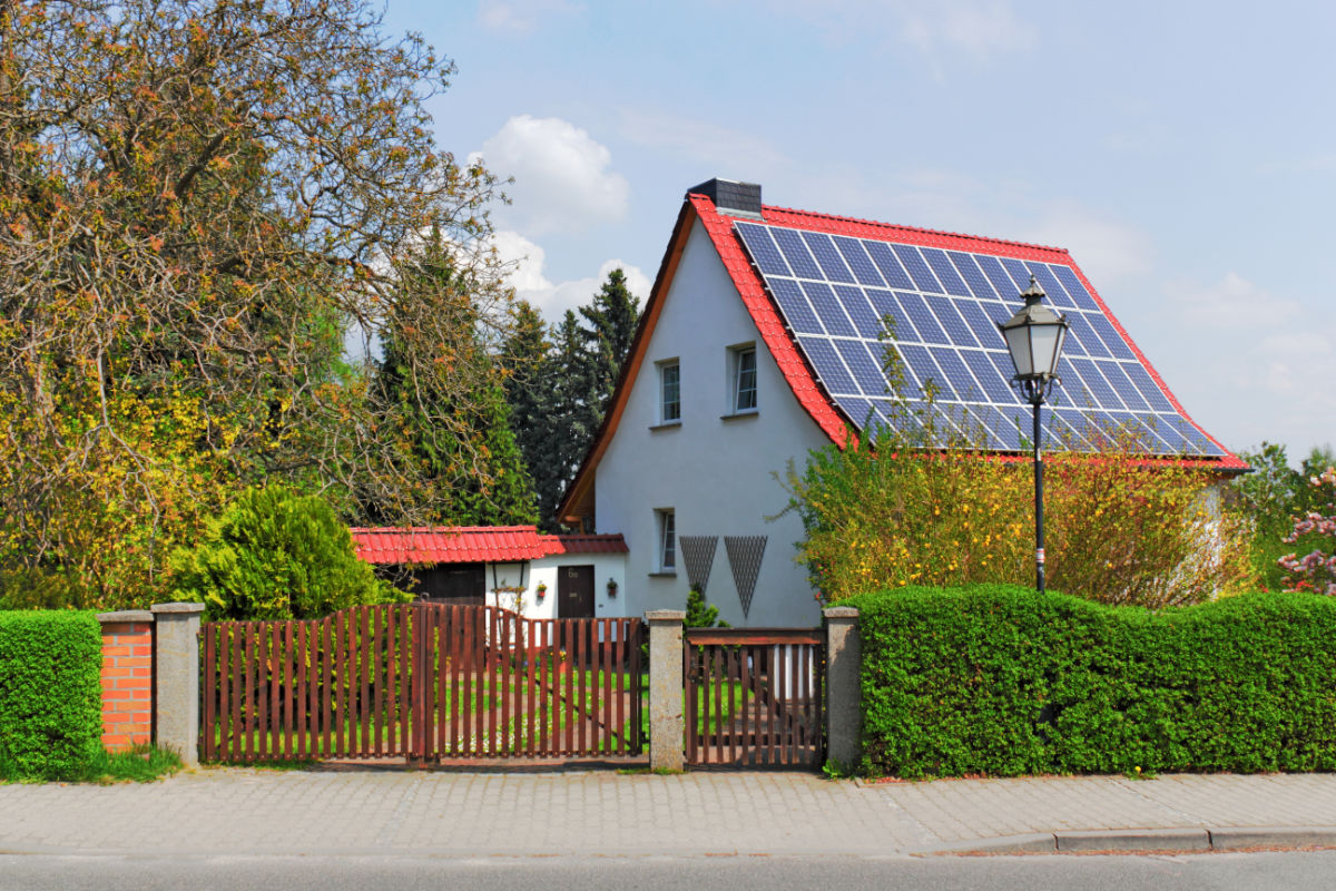 Einfamilienhaus mit einem roten Dach, das mit Solarpaneelen für Solarenergie bedeckt ist, umgeben von grünen Büschen und einem Holzzaun.