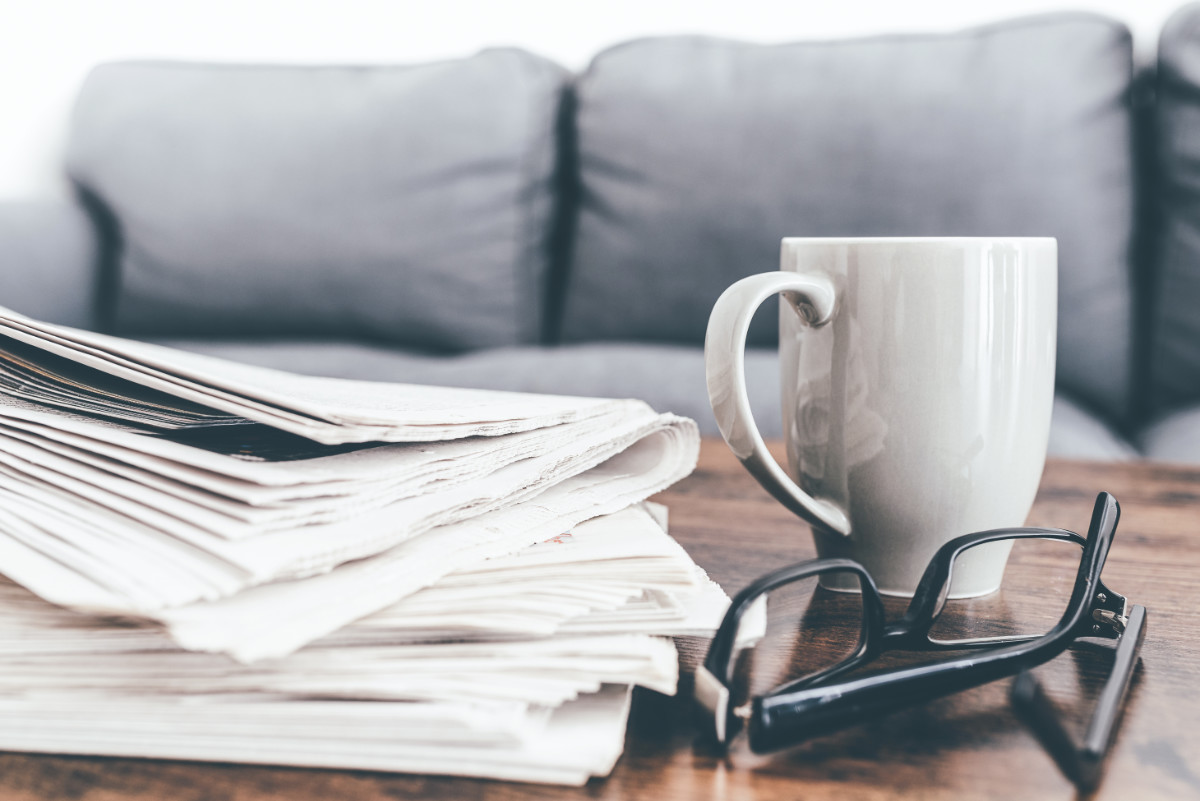 Auf einem Holztisch liegen gefaltete Zeitungen, daneben eine graue Tasse und eine schwarze Brille vor einem verschwommenen grauen Sofa.