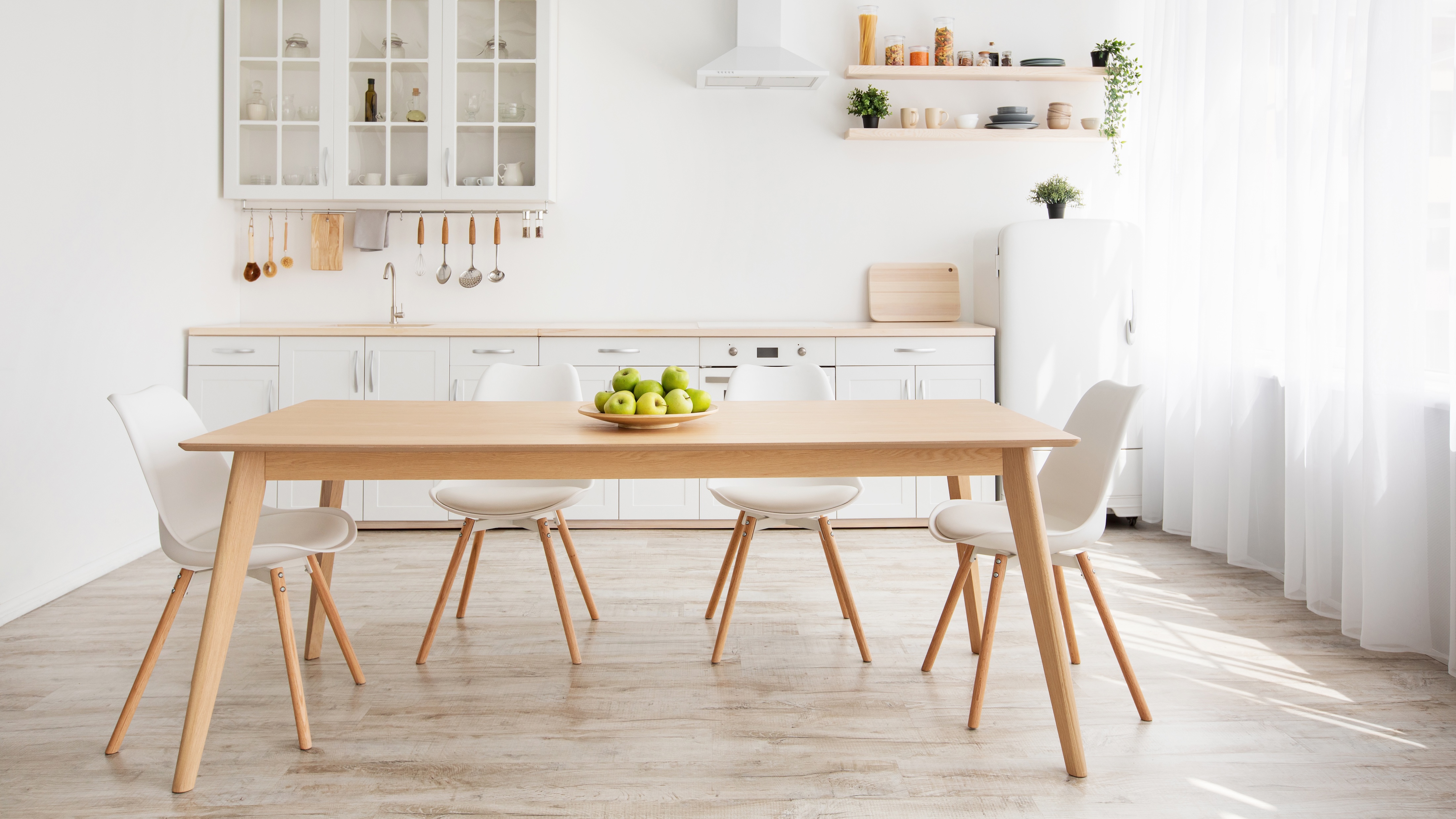 Moderner, minimalistischer Küchenbereich mit weißer Küchenzeile, Holzarbeitsplatte, freistehendem Holz-Esstisch mit vier weißen Stühlen, Obstschale mit grünen Äpfeln in der Mitte und großem Fenster mit weißen Gardinen rechts.