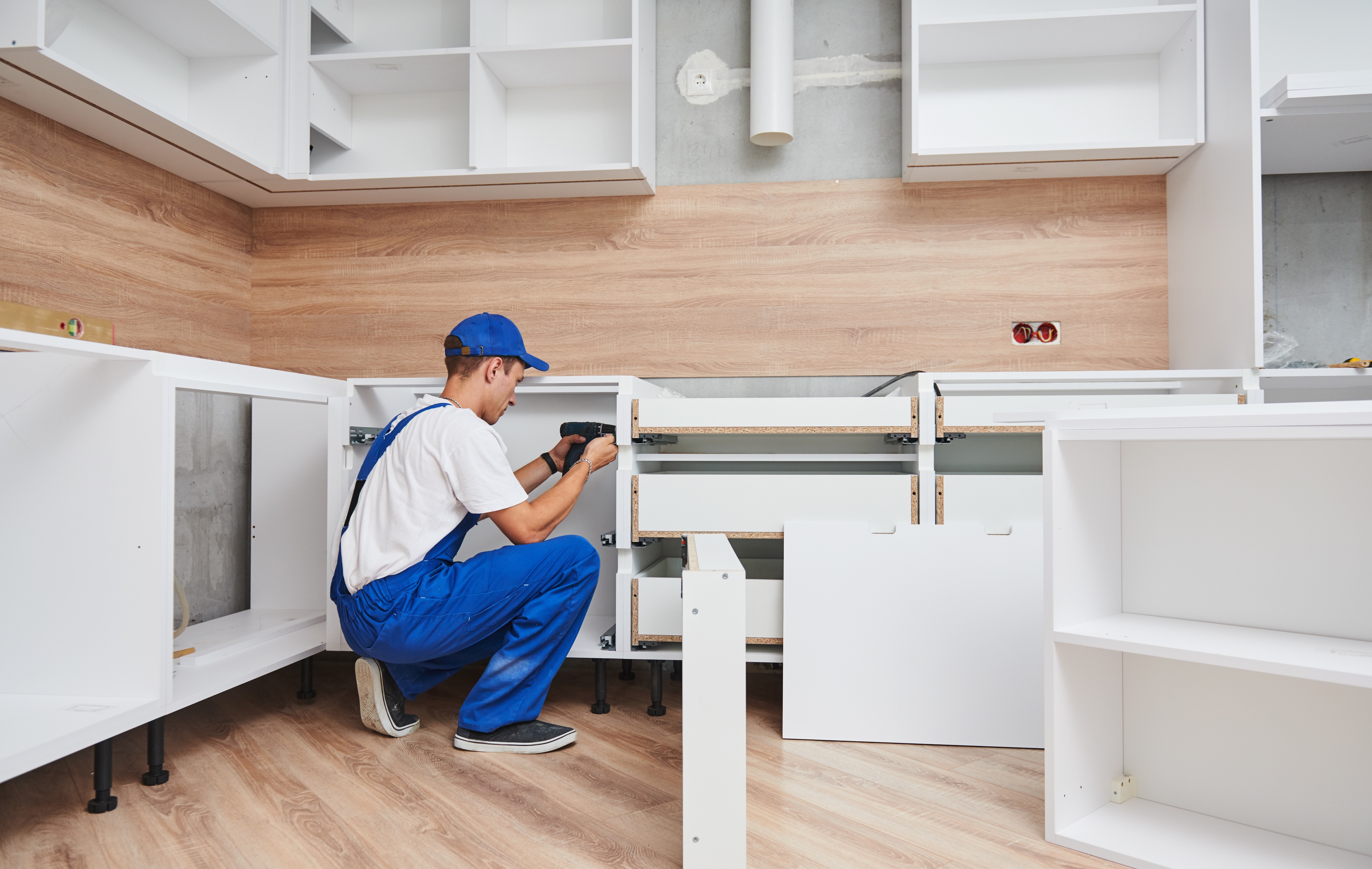 Ein Handwerker in blauer Latzhose und weißem T-Shirt montiert Küchenschränke in einer modernen, unfertigen weißen Küche mit hellbraunem Holzfußboden und Rückwand.