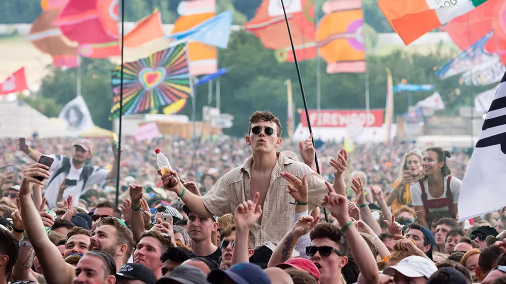 A young man is on the shoulders of someone in a crowd at a festival, with people behind him holding flags