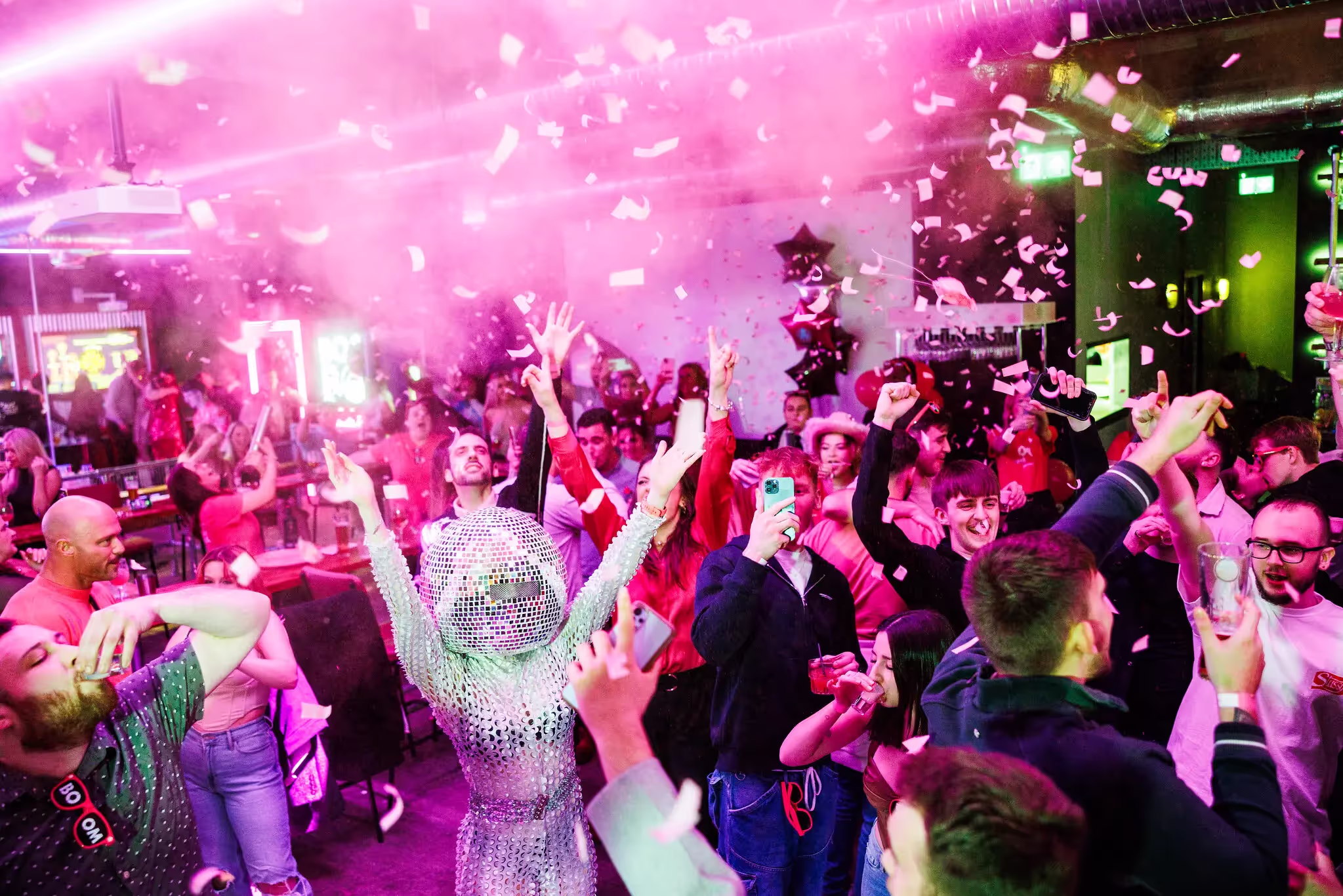 A crowd of people are drinking and dancing under an explosion of pink confetti. One person is dressed like a glitterball