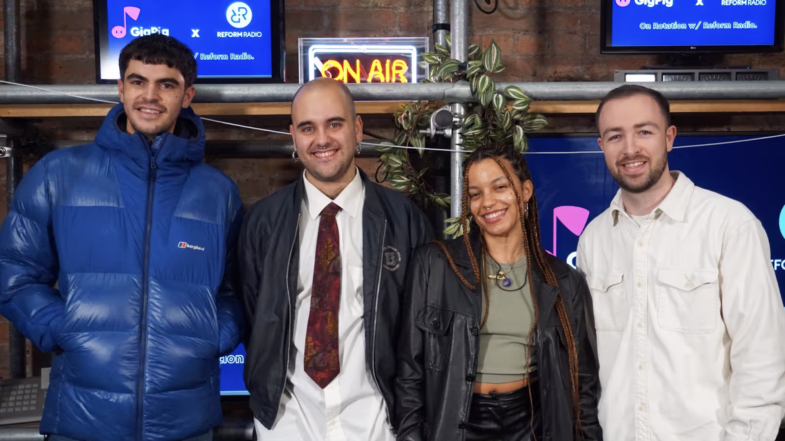 A group of four people smile at the camera with a GigPig screen in the background as well as an ON AIR sign