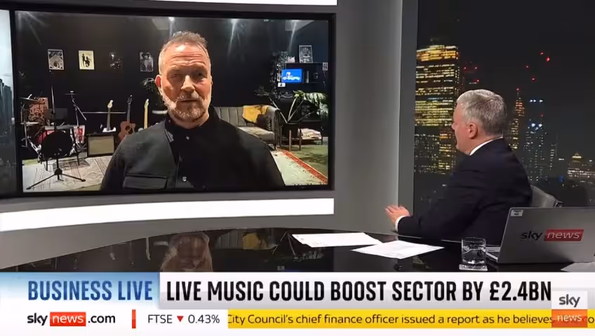 A man sits at a desk in a TV studio, interviewing a bearded man who appears on a television screen