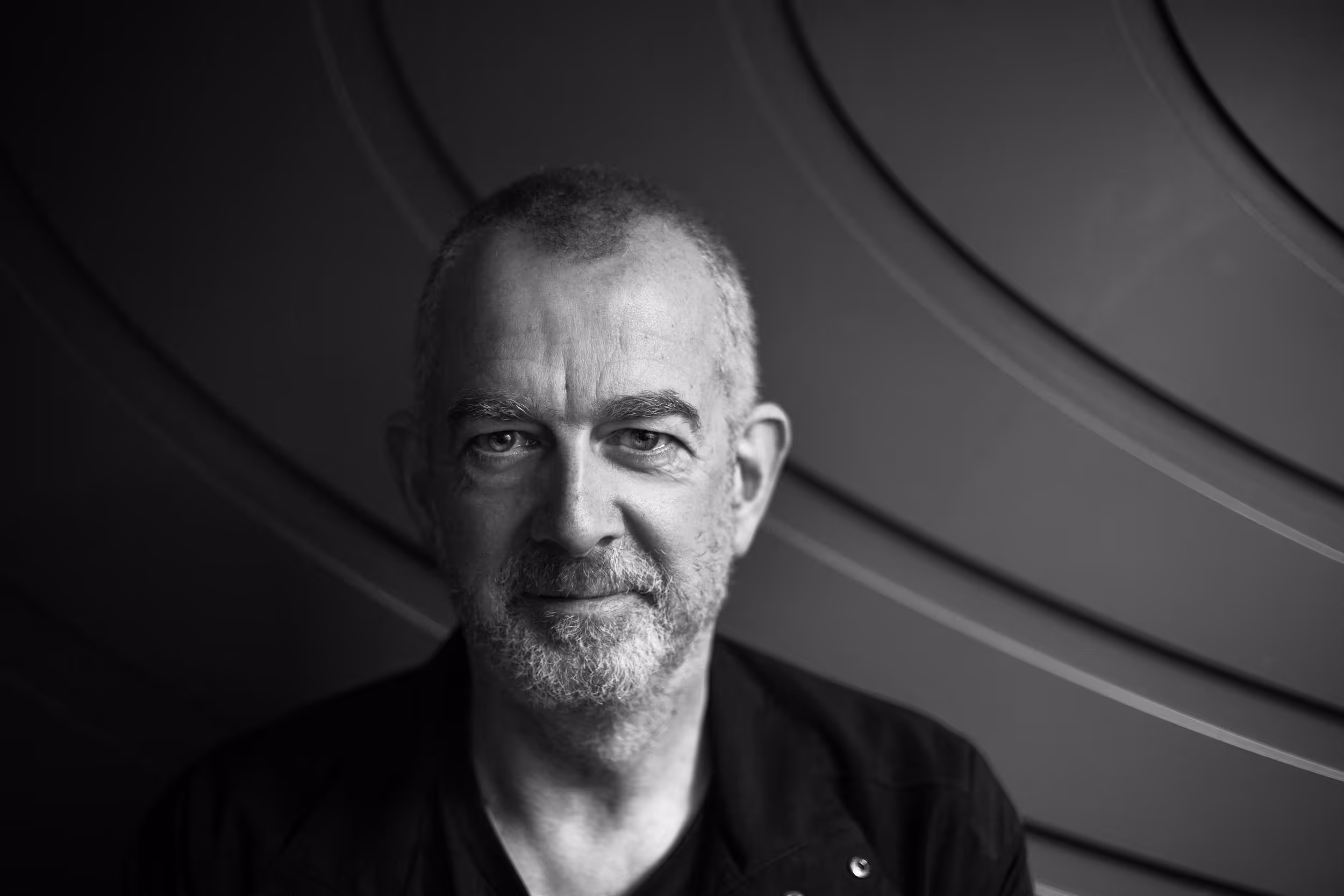 A shot of a man looking into the camera in a serious manner wearing a black jacket and khaki cap