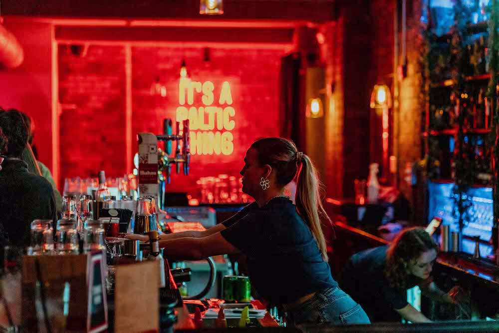 A woman wearing denim is behind a bar reaching over to gather glasses in a dimly lit bar