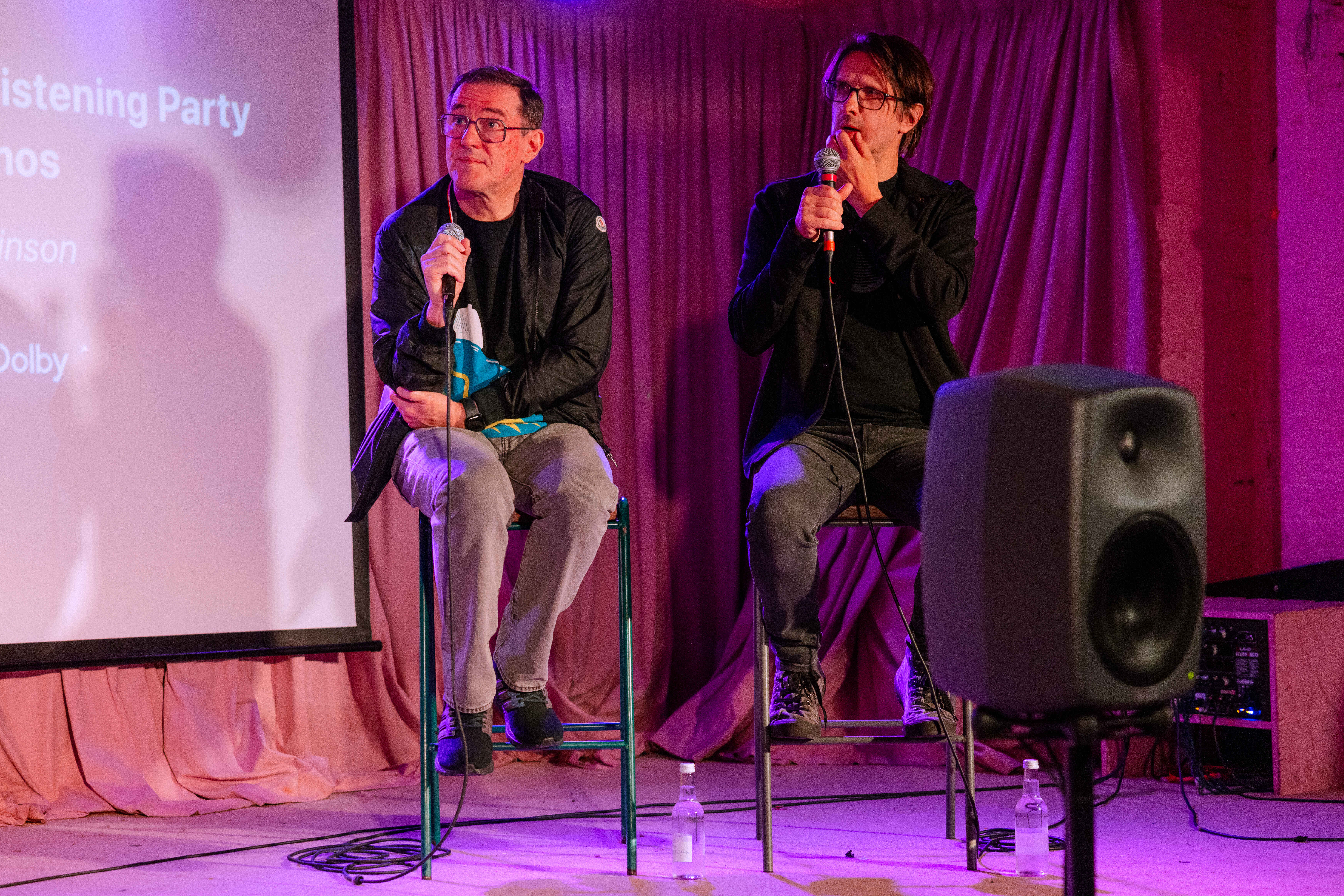 Two men are on a stage looking engaged in conversation, with a speaker and projector screen behind them