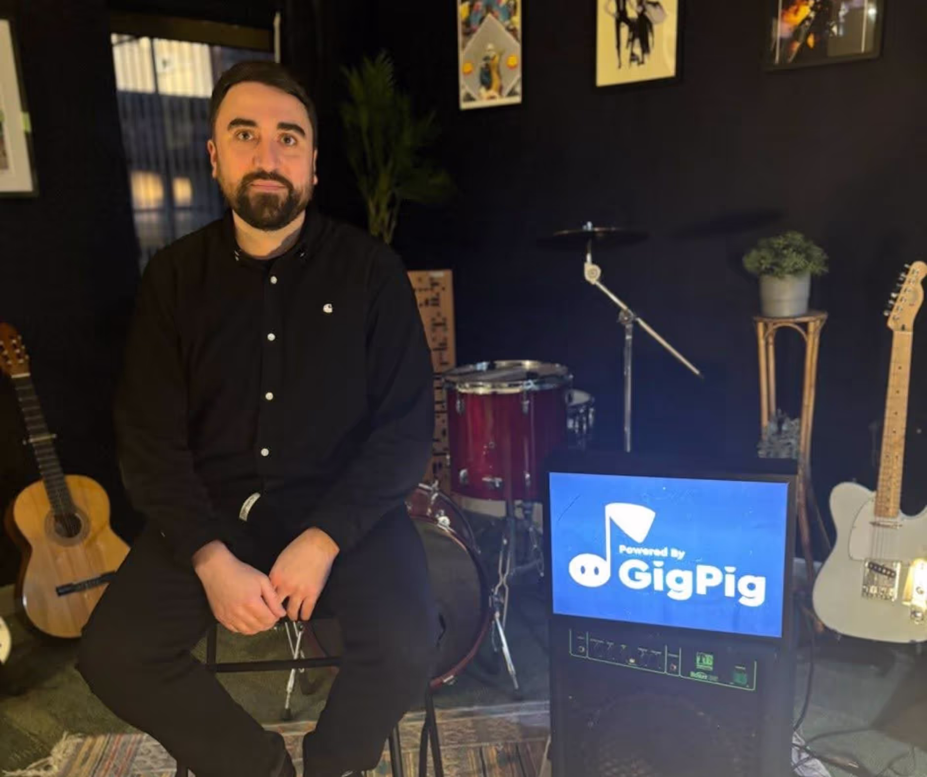 A bearded man dressed in black sits amongst various musical instruments and a GigPig sign
