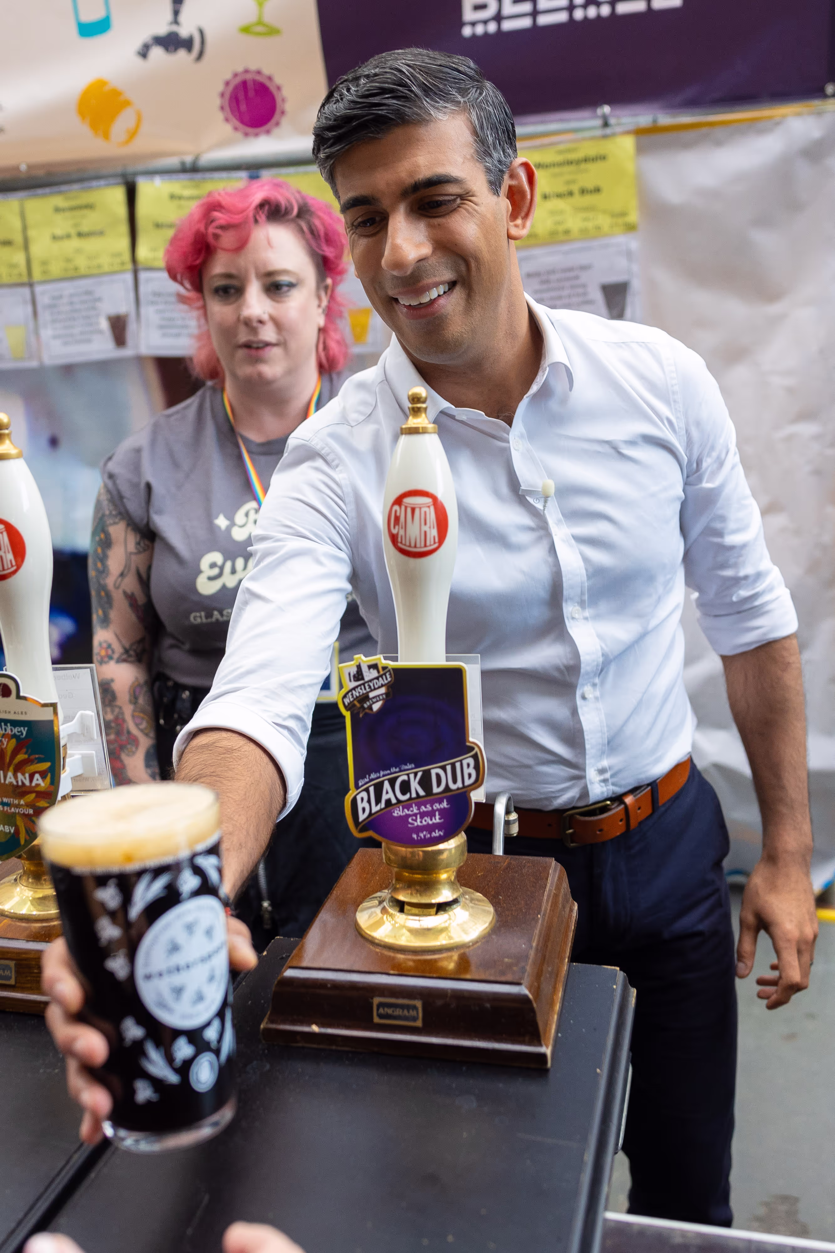 Image of Rishi Sunak smiling into the camera while holding a bar cloth that says "Beer is the answer"