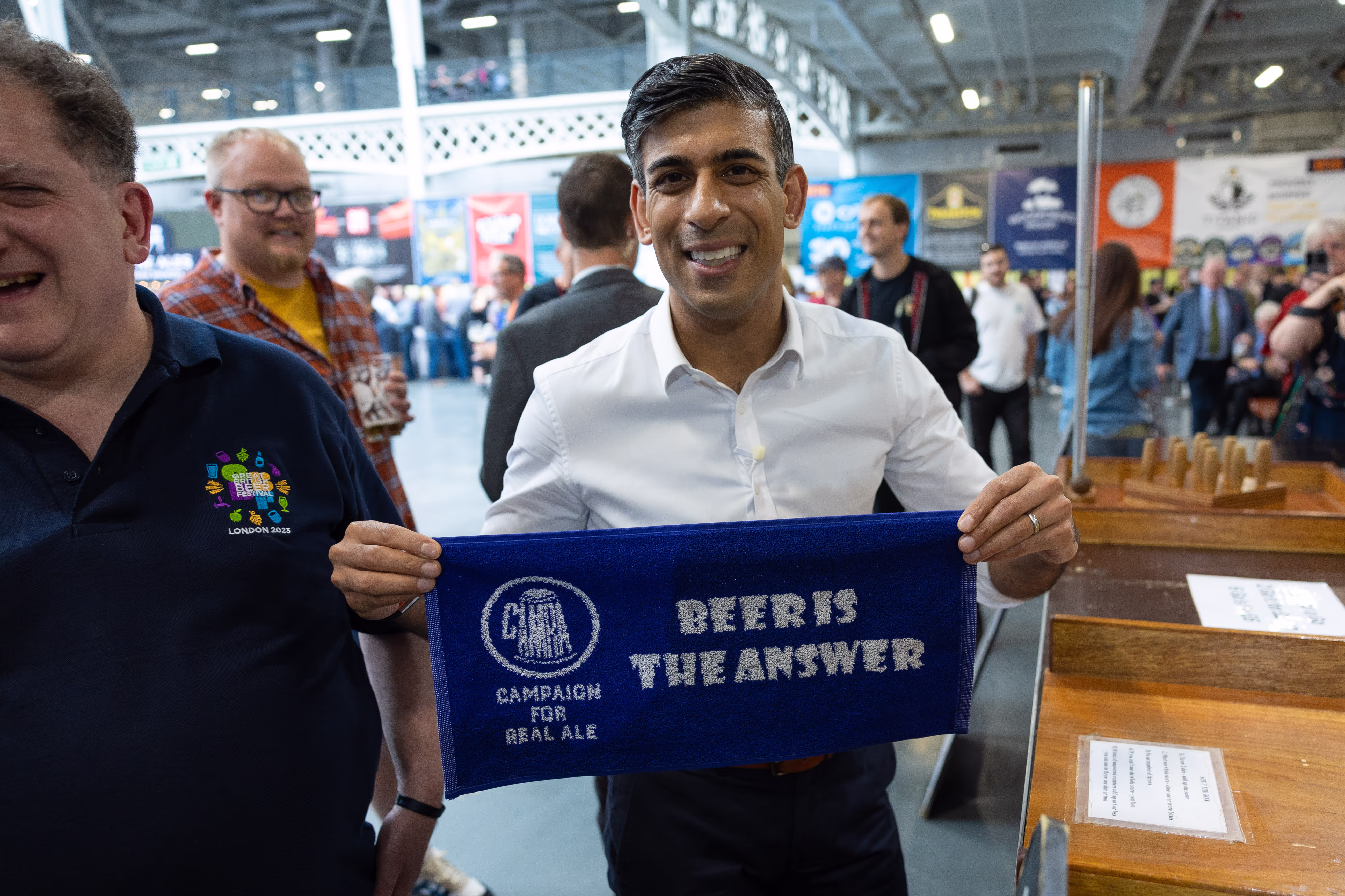 Image of Rishi Sunak smiling into the camera while holding a bar cloth that says "Beer is the answer"