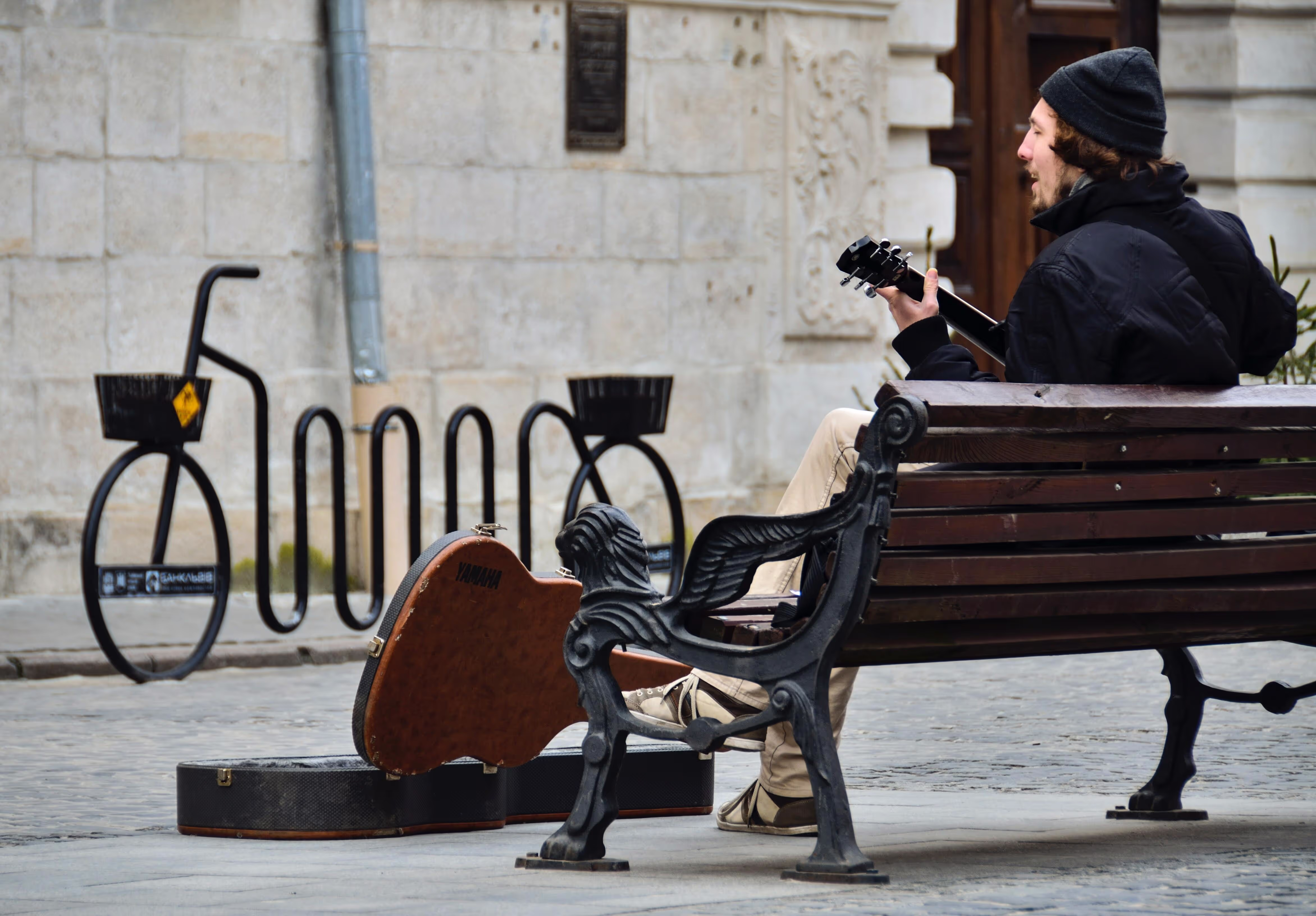 A man sits on a chair outside with a guitar in his lap singing with a guitar case at his feet