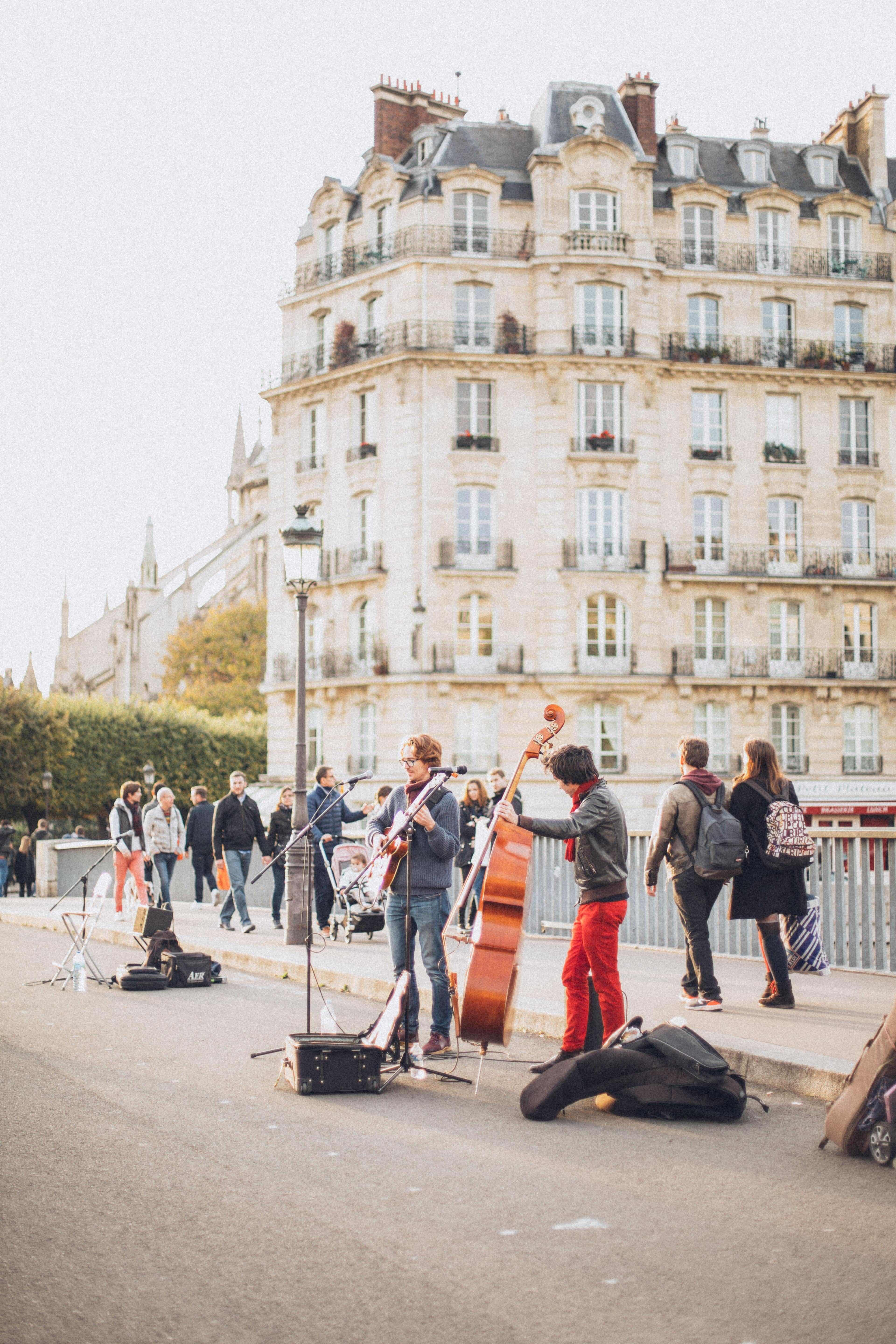 A man sits on a chair outside with a guitar in his lap singing with a guitar case at his feet 